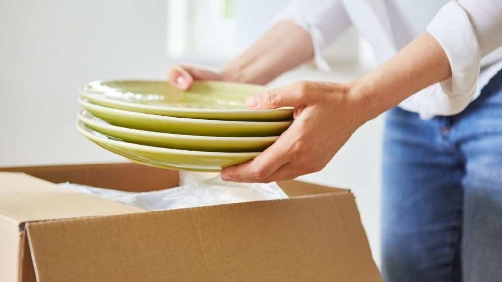 A woman holding a stack of green plates next to a packing box. 